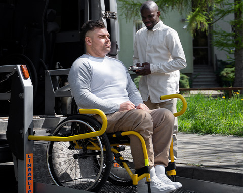 A man in a wheelchair is supported by a companion while getting ready to board a van for transport support