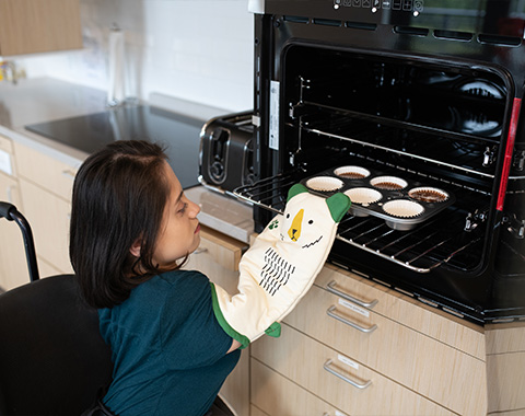 A woman in a wheelchair places muffins into an oven, showcasing her independence for Lifestyle support