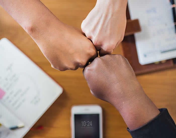 Four hands of diverse skin tones fist-bumping over a cluttered wooden table containing a notebook, papers, and a smartphone. The hands form a circle, symbolizing unity and teamwork.